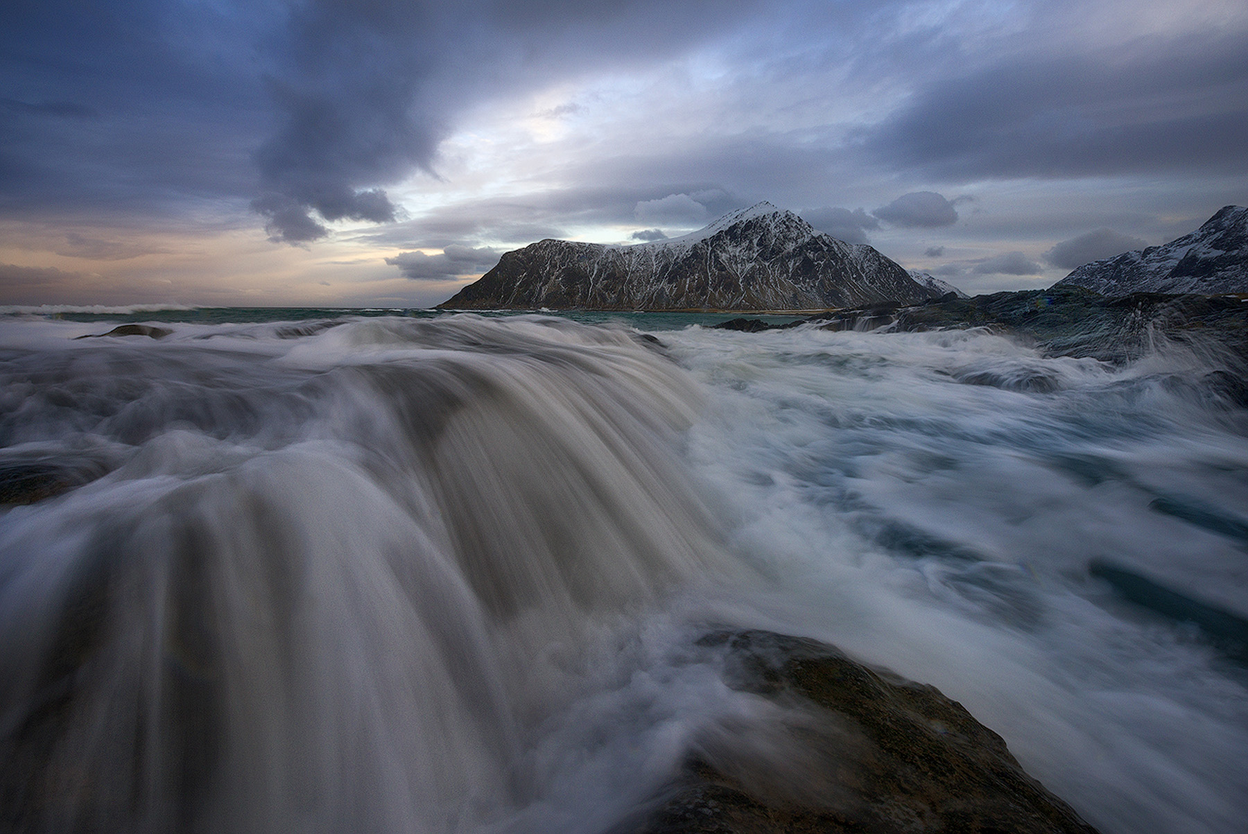 Long exposure of waves washing over a rocky shore below snowy mountains in Lofoten, Norway, shot on a tripod during a winter photography workshop