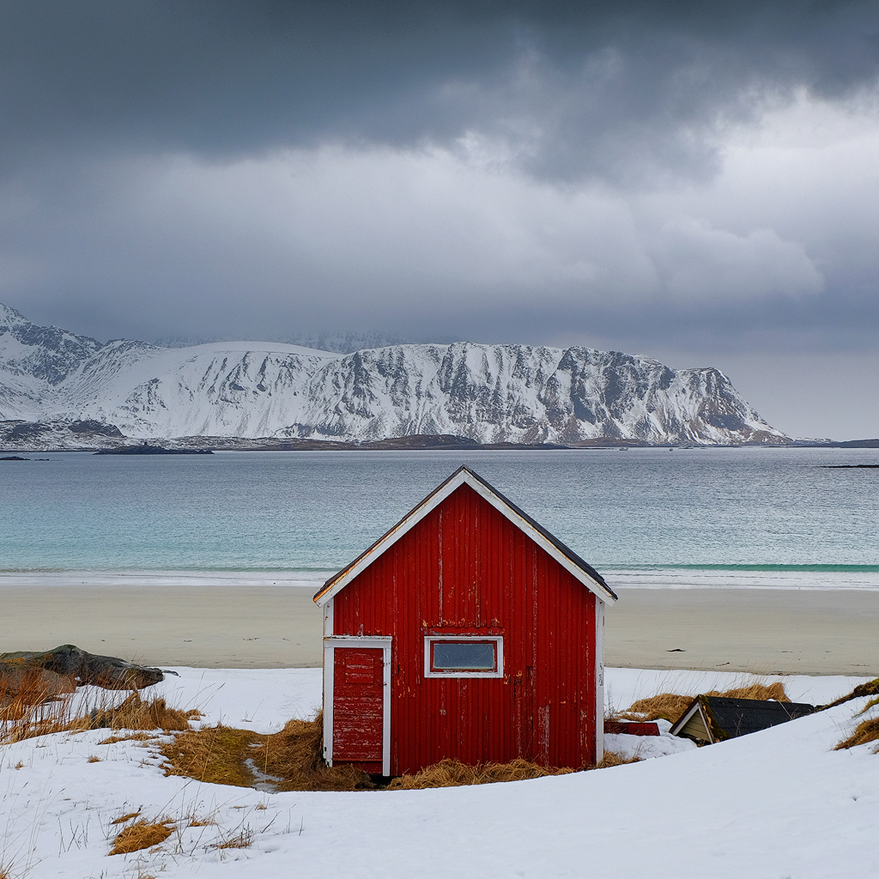 Red cabin on a snow-covered beach below Lofoten mountains under a gray winter sky