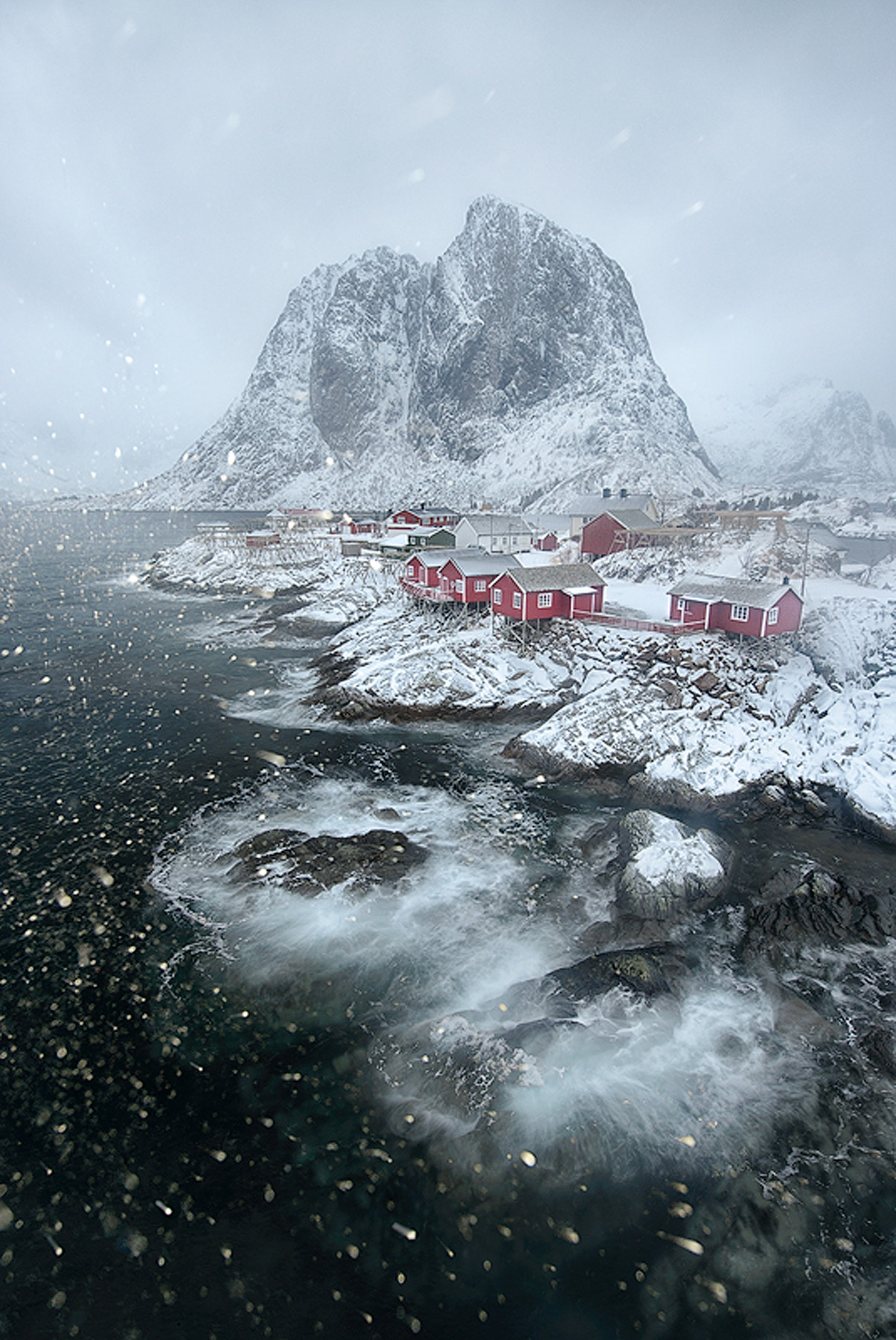 Snow falling over a Lofoten fishing village below a snow-covered mountain in winter