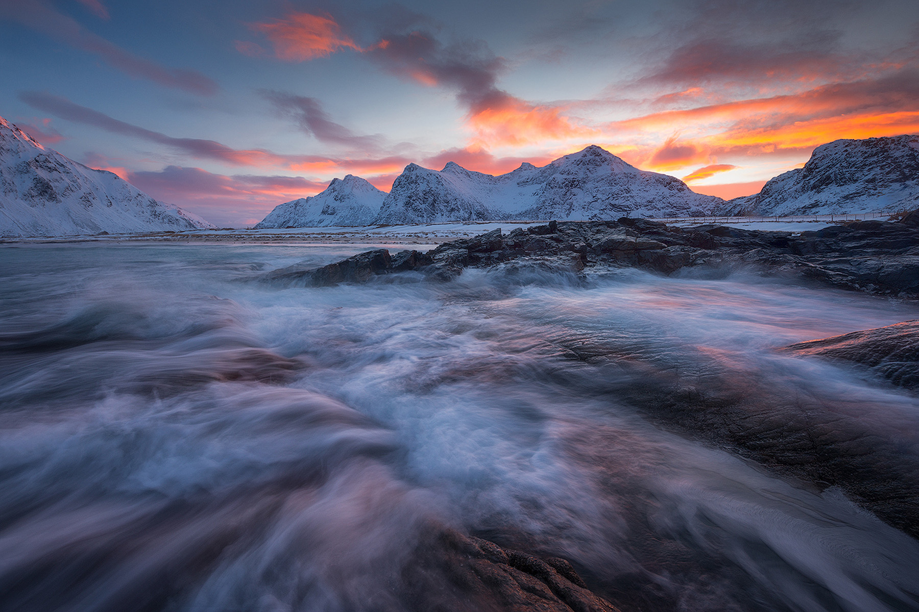 Long exposure of waves washing over rocks under a pink winter sunset with snow-covered peaks in Lofoten, Norway