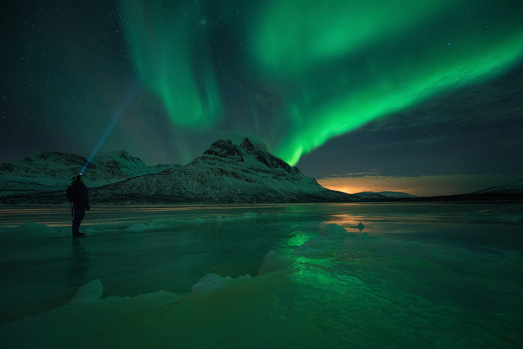 Photographer standing on a Lofoten beach under a green arc of northern lights during a winter photography workshop in Norway