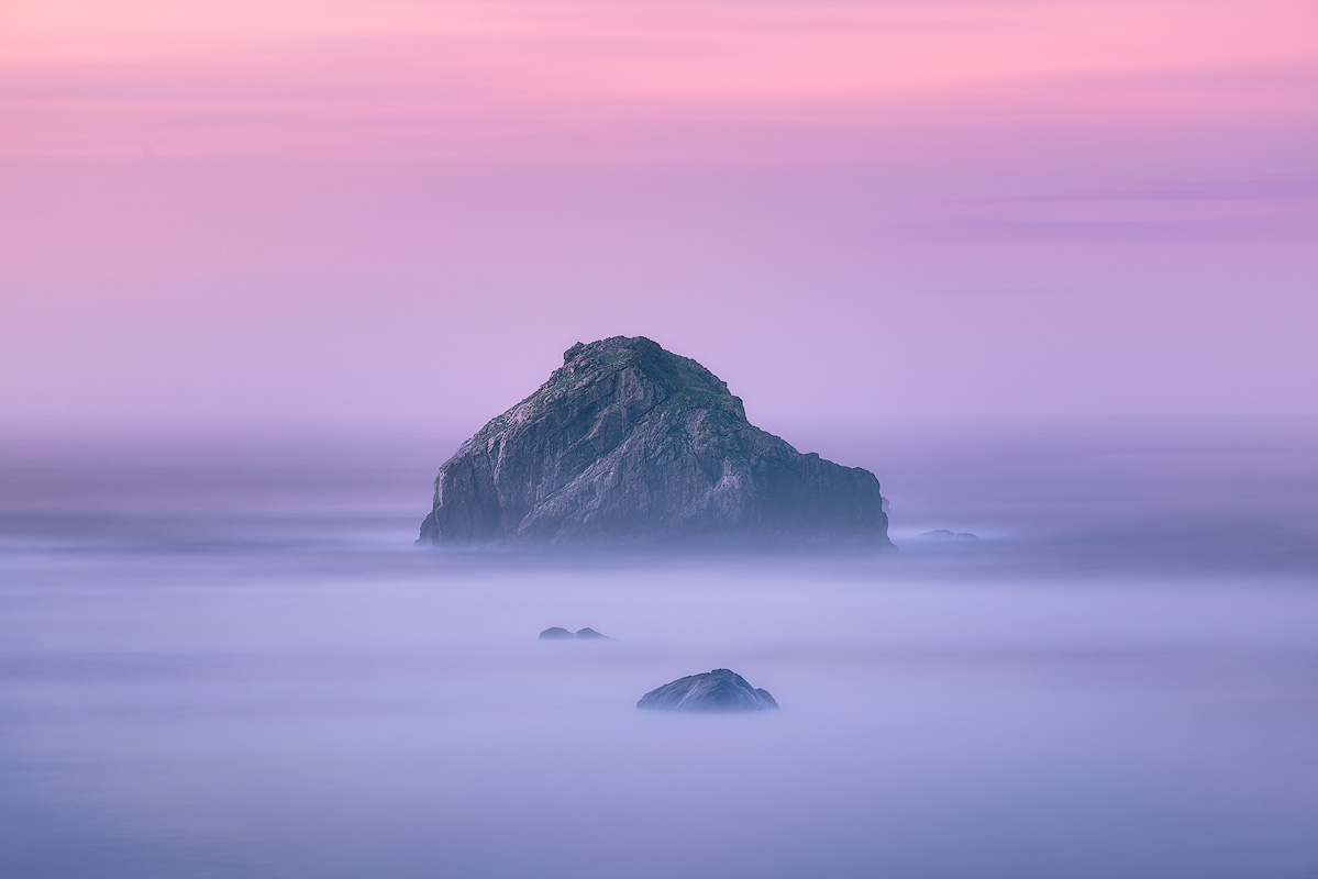 Sea stack rising from silky smooth long exposure ocean in lavender twilight demonstrating ND filter technique that transforms water into fog