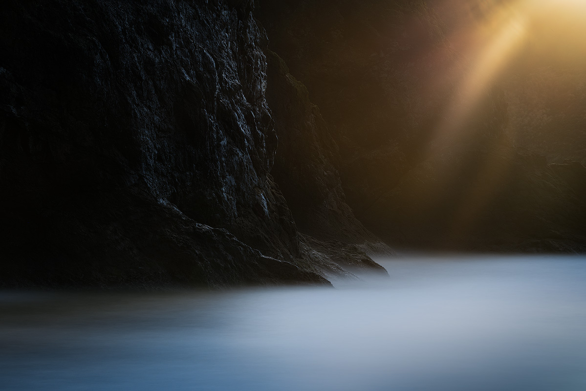 Long exposure photograph of a dark coastal cliff with golden light rays streaming across silky smooth ocean water at Cape Disappointment demonstrating slow shutter speed technique