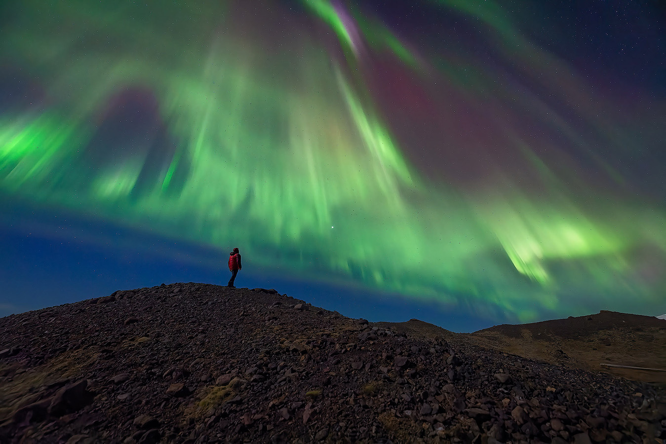 Photographer Michael Schultz standing on a rocky ridge beneath a powerful aurora borealis display with an overhead corona