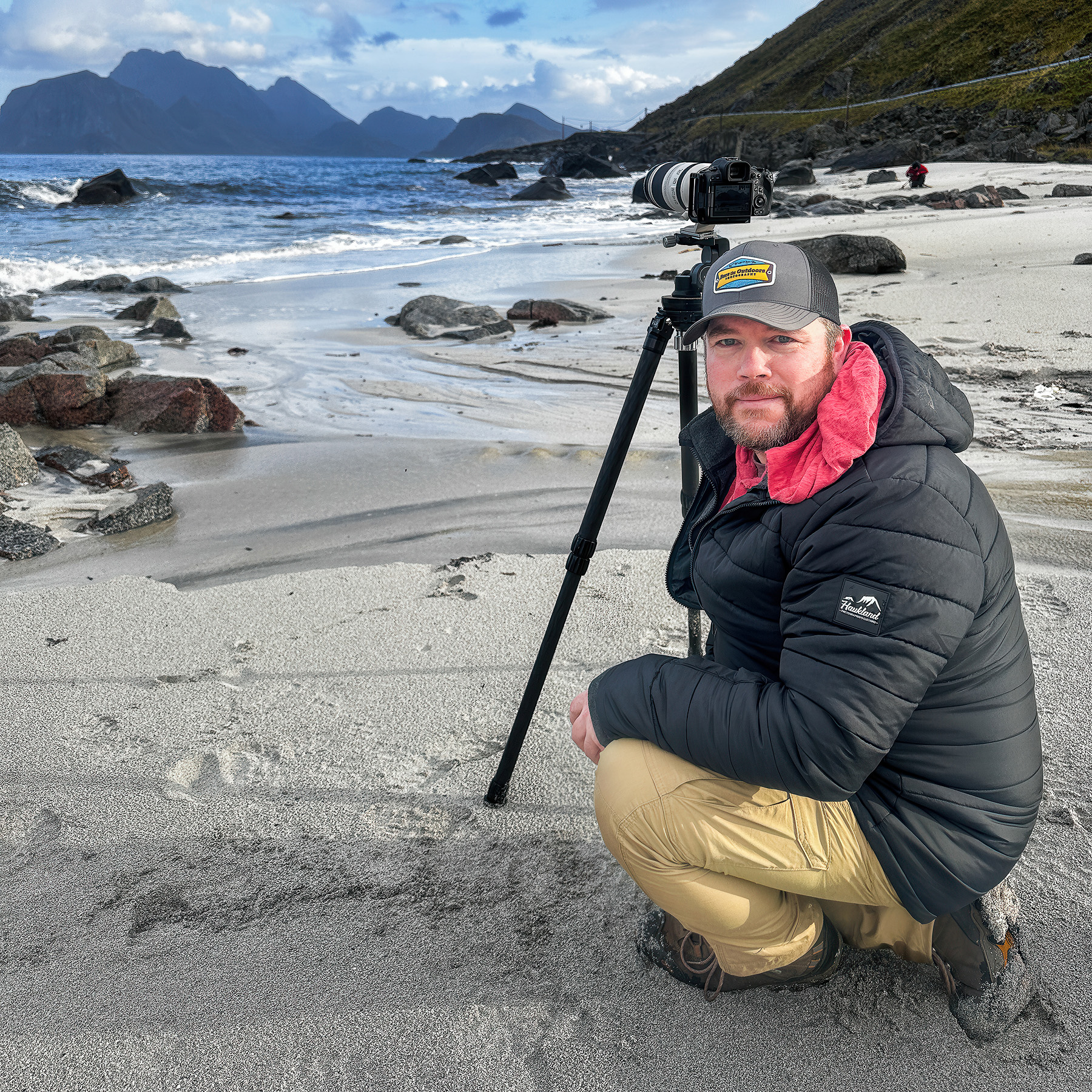 Michael Schultz, Lofoten photography workshop instructor, kneeling on a beach in Lofoten, Norway next to his camera and tripod with the sea and mountains behind him