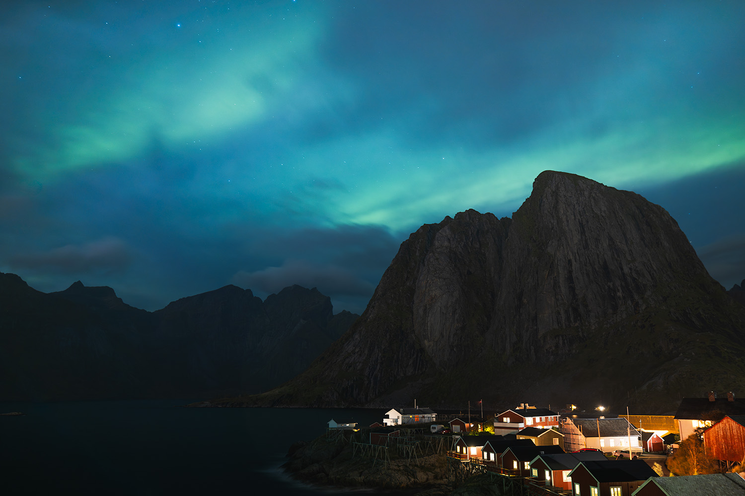 Northern lights dance in the sky behind clouds over cabins and mountains in Hamnoy, Norway