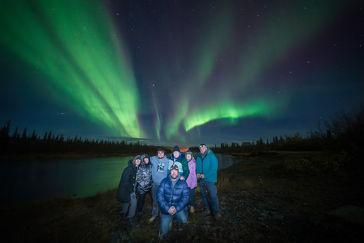 Face The Outdoors Photography workshop group posing under vibrant green aurora borealis in Alaska demonstrating outdoor photography community and shared passion