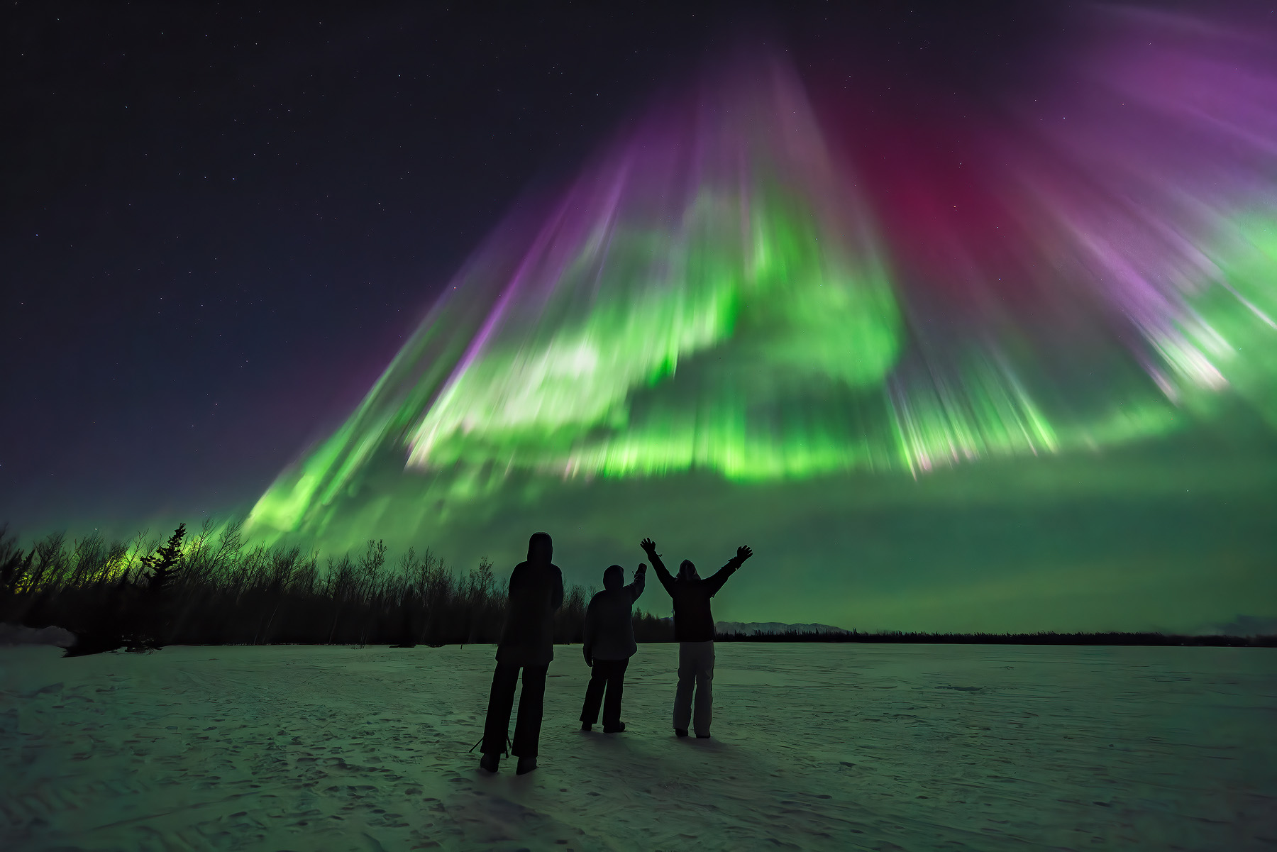 Three people silhouetted on frozen lake with arms raised in awe as massive green and purple aurora borealis corona explodes directly overhead with radiating curtains during intense geomagnetic storm