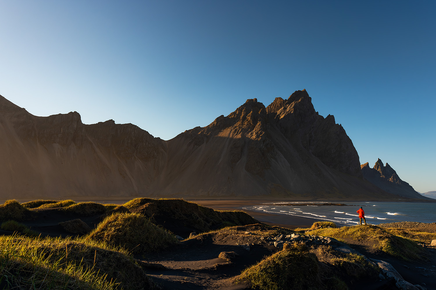 A person in a red jacket standing near a beach with mountains in the background during sunset at Vestrahorn Iceland.