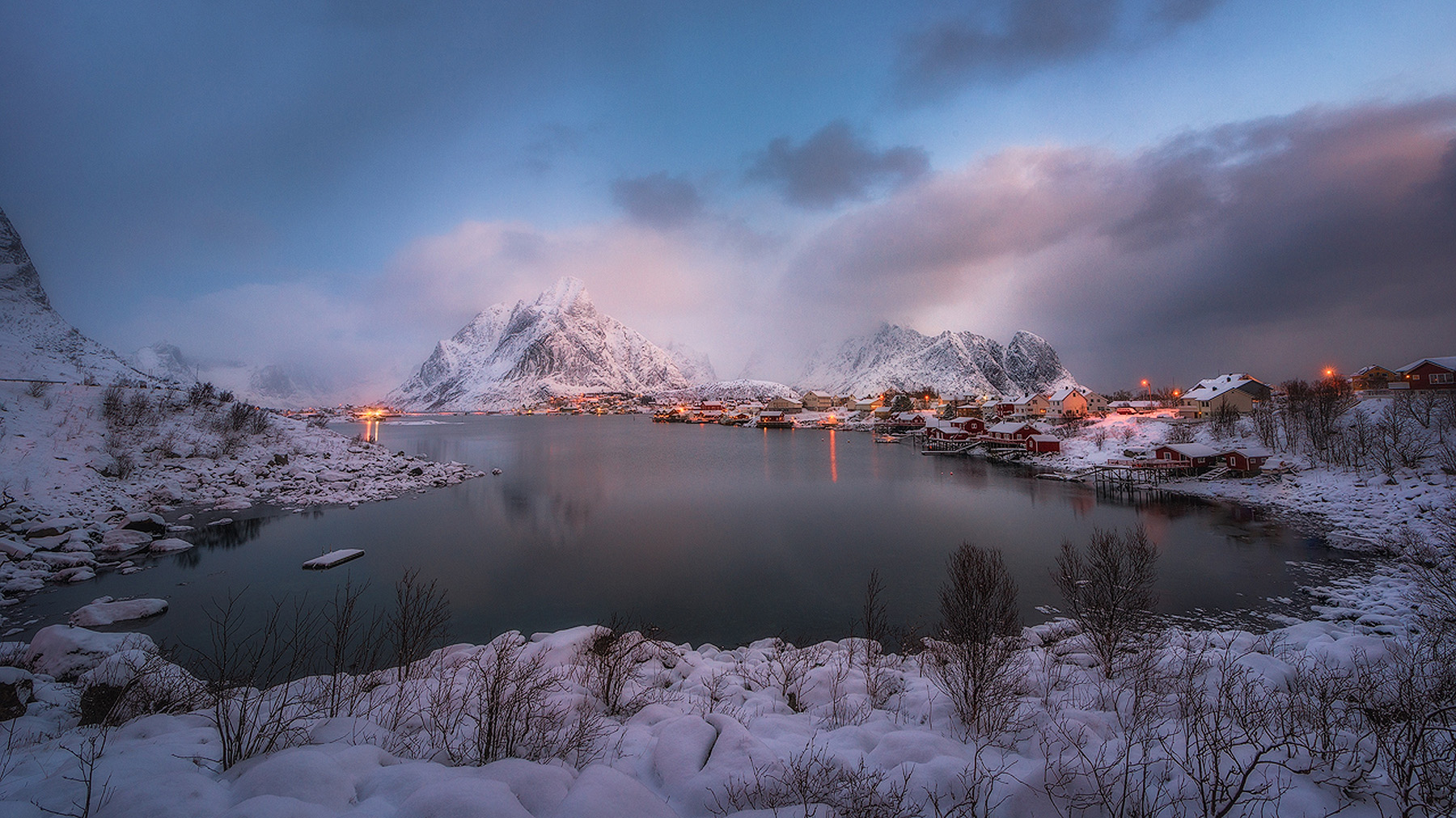 Reine fishing village in Lofoten at dawn in winter, with snow-covered peaks, glowing cabin lights and a calm fjord