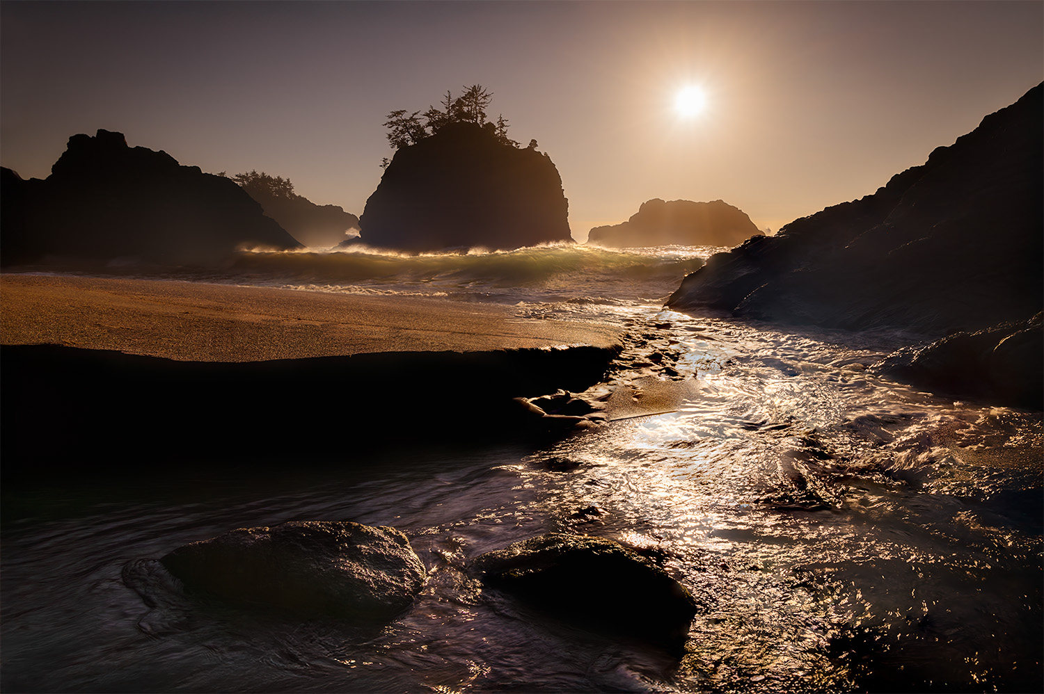A feeder creek flows to the incoming tide, with sunset light illuminating the waves and casting a warm glow on the sandy beach and sea stacks.