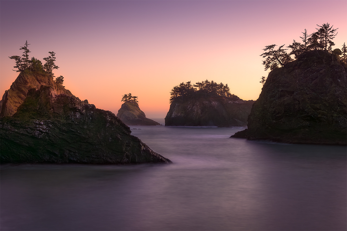 Four sea stacks glow in the warm light of sunset, with a long exposure softening the rough surf, creating a peaceful coastal scene.